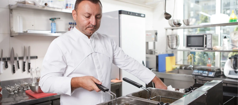 individual in a chef's uniform working in a commercial kitchen. The chef is focused on cooking or preparing food using a deep fryer. The kitchen is equipped with various cooking utensils and appliances, such as knives set on a magnetic rack, a microwave, and other culinary tools. It's a well-lit space, suggesting a professional environment for food preparation.