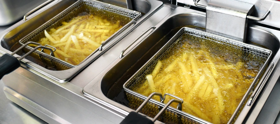 wide-angle view of two baskets full of French fries being deep-fried in a professional kitchen setting. Each basket is inside its own section of a commercial deep fryer, and we can see the oil actively bubbling around the fries, indicating that they are being cooked. The equipment is made of stainless steel, which is typical for food service due to its durability and ease of cleaning.