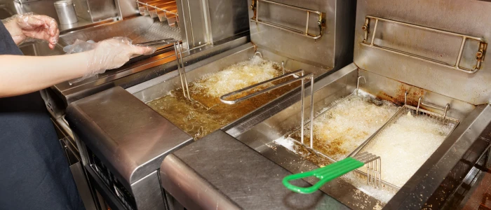 The image shows a commercial kitchen setting with a person using tongs to handle food in a bubbling deep fryer.