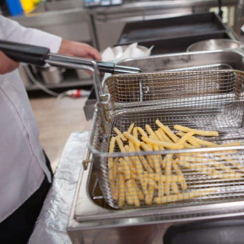 This picture shows a person in a kitchen lifting a basket of freshly fried French fries from a deep fryer. The setup suggests a professional or commercial cooking environment.