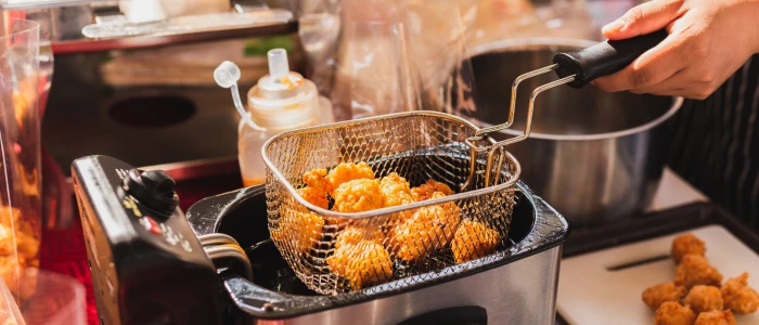 The image displays a hand lifting a basket out of a small deep fryer, filled with freshly fried food, likely chicken or a similar breaded item.