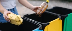 The image shows a person holding an empty plastic bottle and a used egg carton, poised to sort them into separate recycling bins