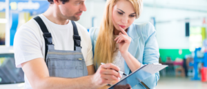 The image depicts two individuals, a man and a woman, focused on a document that the man is holding. The man appears to be wearing a white shirt with overalls, which is typical attire for manual work such as maintenance or technical services. This suggests he might be a technician or mechanic. The woman is wearing a light blue blazer and a white lace top, which suggests a more formal or business-oriented role, possibly a manager or client. They are both looking at the document with attentive expressions, indicating they are discussing something important, likely related to the man's work or the services provided. The background is blurred, but it seems to be an indoor setting with bright lighting, which could be a workshop, factory floor, or a service center.