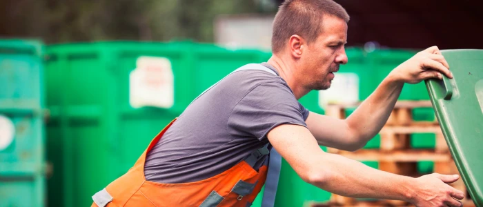 A man in a gray t-shirt and orange work suspenders is actively engaged in recycling tasks, lifting the lid of a green bin at a recycling center with other green containers in the background, indicating a focus on environmental responsibility.