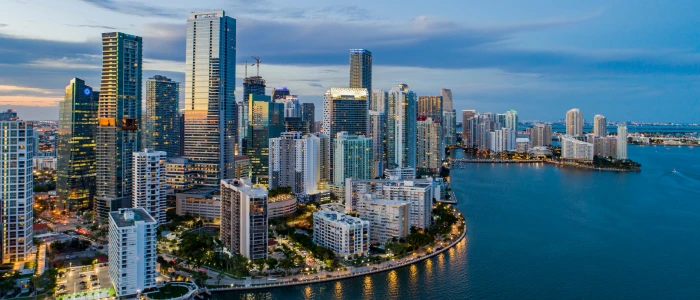 The image showcases a stunning aerial view of a dense urban skyline at twilight, with numerous skyscrapers clustered close to the waterfront. This is indicative of a modern cityscape, likely that of a major city known for its high-rise buildings. The city is situated on a waterfront, suggesting a location that is either coastal or along a large river or bay. The buildings are illuminated, reflecting the city's active nightlife and commercial areas. The water in the foreground adds a calming element to the bustling city vibe. This could be a scene from a well-known city like Miami, known for its picturesque skyline along Biscayne Bay.