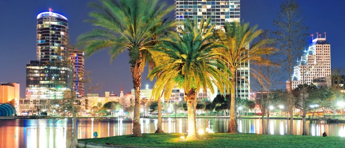 A panoramic night view of downtown Orlando with illuminated high-rise buildings reflecting on a lake, foregrounded by vibrant palm trees. The cityscape exudes a tranquil yet vibrant atmosphere, showcasing urban life alongside natural beauty.